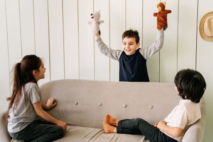 Três crianças assistem a uma apresentação de fantoches em um ambiente interno e acolhedor.