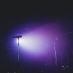 Microfone em um palco vazio iluminado por luzes roxas, preparado para uma apresentação musical.