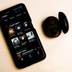 Smartphone exibindo uma interface de aplicativo de música ao lado de fones de ouvido sem fio sobre fundo neutro