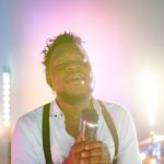 Cantor negro se apresenta ao microfone em um palco com estética retrô.
