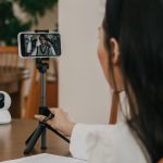 Mulher faz uma videochamada pelo smartphone apoiado em um suporte, em ambiente interno.