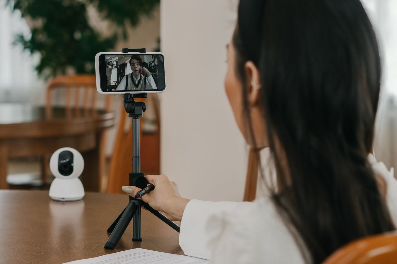 Mulher faz uma videochamada pelo smartphone apoiado em um suporte, em ambiente interno.