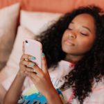 Mulher de cabelos cacheados usa um smartphone enquanto exibe unhas coloridas.