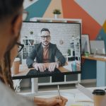 Equipe participa de uma videoconferência com um colega exibido na tela de um computador em um escritório colorido.