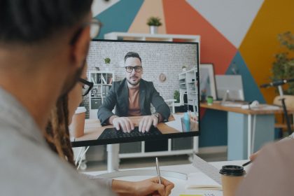 Equipe participa de uma videoconferência com um colega exibido na tela de um computador em um escritório colorido.