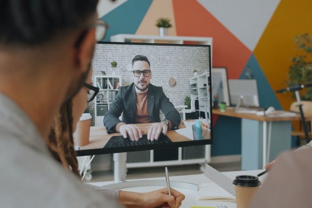 Equipe participa de uma videoconferência com um colega exibido na tela de um computador em um escritório colorido.