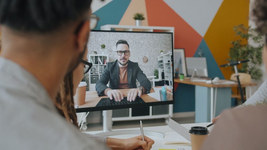 Equipe participa de uma videoconferência com um colega exibido na tela de um computador em um escritório colorido.
